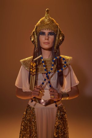 Stilvolle Frau mit ägyptischem Kopfschmuck und Blick, die Gauner und Dreschflegel auf braunem Hintergrund hält