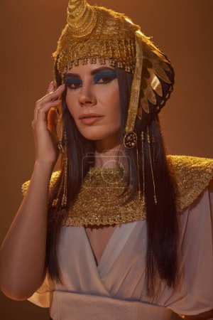 Retrato de mujer morena en aspecto egipcio tradicional y maquillaje audaz de pie aislado en marrón