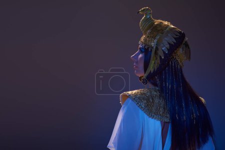 Foto de Vista lateral de la mujer con estilo en tocado egipcio de pie sobre fondo marrón con luz azul - Imagen libre de derechos