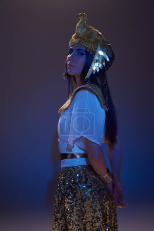 Mujer elegante en aspecto egipcio dorado y tocado mirando a la cámara en marrón con luz azul