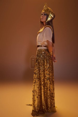 Volle Länge der Frau in goldenem ägyptischen Look und Kopfbedeckung auf braunem Hintergrund