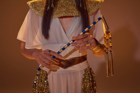 Ausgeschnittene Ansicht einer Frau im ägyptischen Outfit, die Dreschflegel hält, während sie auf braunem Hintergrund posiert