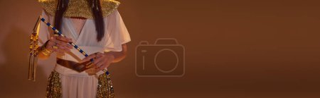 Foto de Vista recortada de la mujer en mirada egipcia elegante celebración de mayal sobre fondo marrón, bandera - Imagen libre de derechos