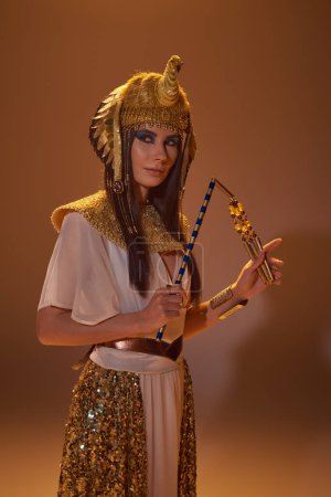 Schöne Frau in ägyptischer Kleidung und Kopfschmuck hält Dreschflegel, während sie auf braunem Hintergrund posiert