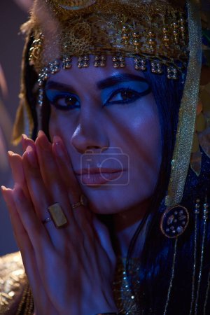 Portrait of woman with makeup and egyptian headdress looking at camera in blue light on brown