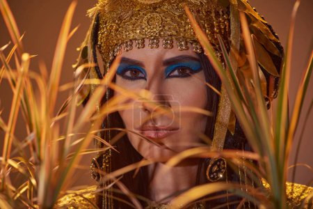Elegant woman in egyptian headdress and bold makeup posing behind desert plants isolated on brown