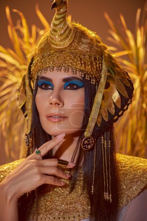 Elegante Frau im ägyptischen Look und Kopfschmuck posiert neben verschwommenen Pflanzen auf braunem Hintergrund
