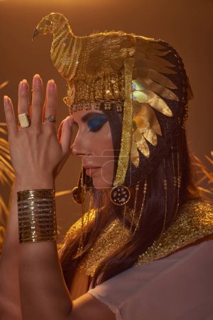 Femme en costume égyptien faisant geste mains de prière près de plantes floues sur fond brun