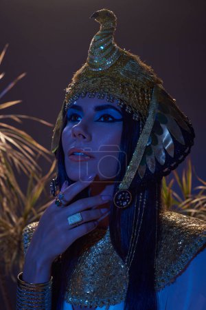 Porträt einer Frau mit ägyptischem Kopfschmuck, die in der Nähe von Pflanzen in blauem Licht auf braunem Hintergrund wegschaut