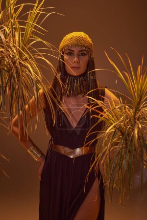 Femme en coiffe égyptienne et collier posant entre les plantes du désert isolé sur brun