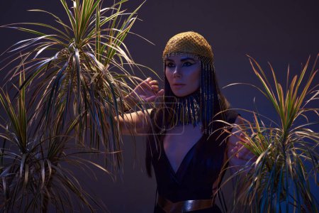 Femme élégante au look égyptien et coiffure près des plantes du désert sur fond brun avec lumière bleue