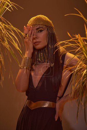 Woman in egyptian outfit and headdress covering face while standing near plants on brown background