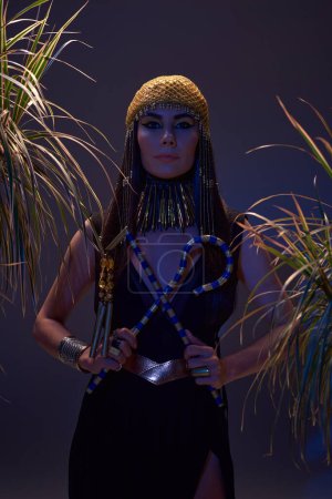 Woman in egyptian look holding flail and crook near plants on brown background with blue light