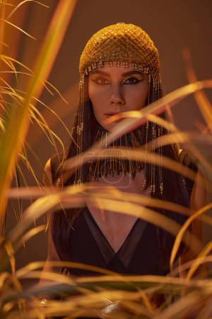 Mujer elegante en estilo egipcio mirando a la cámara cerca de plantas borrosas aisladas en marrón