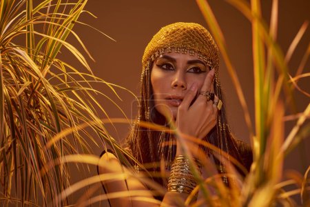 Woman in egyptian headdress and necklace touching cheek near plants isolated on brown