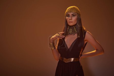 Trendy Frau in ägyptischem Look und Halskette hält Hand auf Hüfte, während sie auf braunem Hintergrund steht