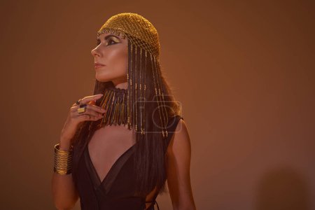 Elegant woman with bold makeup and egyptian costume standing on brown background with light
