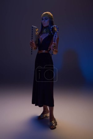 Volle Länge der Frau im ägyptischen Look hält Dreschflegel und Gabel und steht auf braun mit blauem Licht