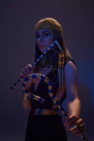 Photo for Stylish woman in egyptian attire holding flail and crook in blue light on brown background - Royalty Free Image