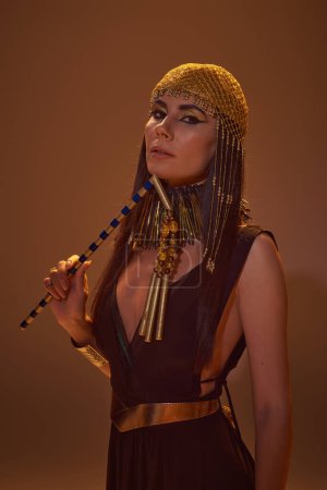 Elegante Frau mit Make-up und ägyptischem Blick hält Dreschflegel in der Hand und blickt in die Kamera auf braunem Hintergrund