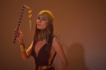Téléchargez les photos : Femme à la mode dans le regard égyptien tenant flair et regardant la caméra sur fond brun - en image libre de droit
