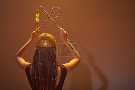Back view of brunette woman in egyptian headdress holding crook and flail on brown background