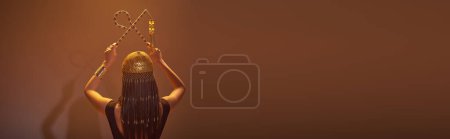 Photo for Back view of woman in egyptian headdress holding crook and flail on brown background, banner - Royalty Free Image