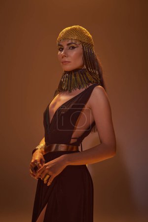 Hermosa mujer morena en traje egipcio y tocado mirando a la cámara sobre fondo marrón