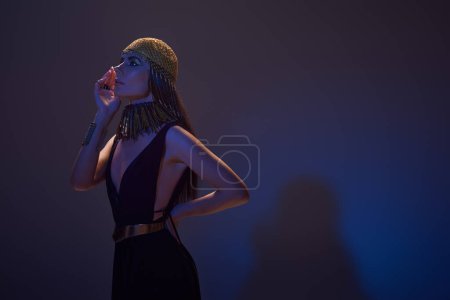 Téléchargez les photos : Modèle élégant en tenue égyptienne et coiffure posant en lumière bleue sur fond violet - en image libre de droit