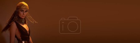 Téléchargez les photos : Femme élégante en robe égyptienne et maquillage regardant la caméra et posant sur fond brun, bannière - en image libre de droit
