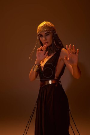 Brünette Frau im ägyptischen Kleid und Blick posiert und schaut in die Kamera isoliert auf braun