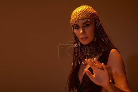 Frau mit ägyptischer Kopfbedeckung und Blick posiert vor Kamera auf braunem Hintergrund mit Licht
