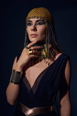 Portrait of woman in egyptian headdress and costume touching chin while standing on blue background