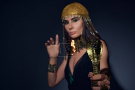 Blurred woman in egyptian look holding crook in snake shape while standing on blue background