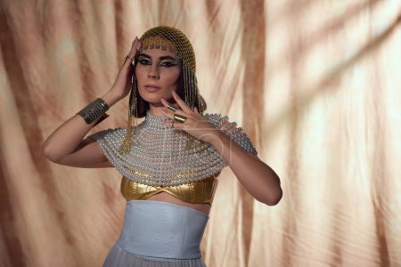 Téléchargez les photos : Femme élégante en coiffure égyptienne et perle haut regardant la caméra sur fond abstrait - en image libre de droit