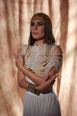 Porträt einer brünetten Frau in Perlentop und ägyptischem Look posiert auf abstraktem Hintergrund