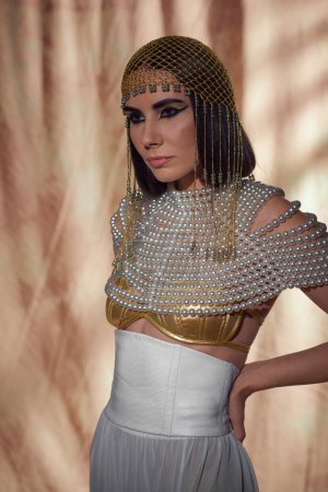 Brünette Frau mit Kopfbedeckung und ägyptischem Look posiert und steht auf abstraktem Hintergrund