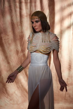 Attraktive Frau mit kühnem Make-up posiert in ägyptischem Kostüm und Perlenoberteil auf abstraktem Hintergrund