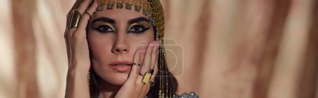 Brunette woman with egyptian makeup and headdress touching face on abstract background, banner