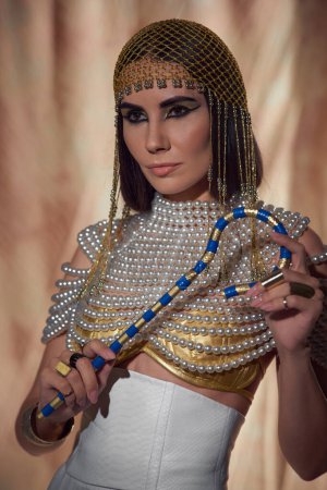 Elegant woman in headdress and pearl top holding egyptian crook on abstract background