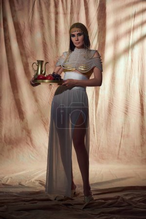 Stylish woman in egyptian outfit holding jug and fruits while posing on abstract background