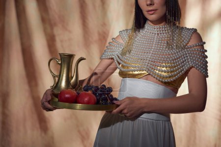 Vista recortada de la mujer en mirada egipcia sosteniendo jarra y frutas mientras posando sobre fondo abstracto