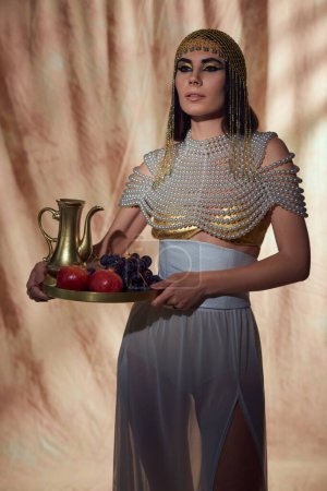 Mujer elegante en aspecto egipcio y la parte superior de perlas que sostiene jarra y frutas sobre fondo abstracto