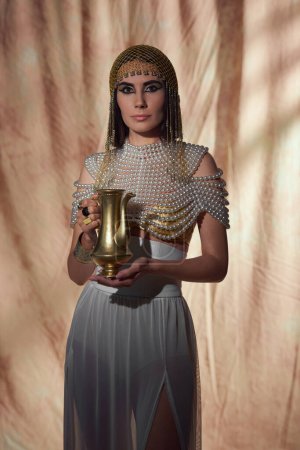 Elegante Frau im ägyptischen Look und Perlenkette mit glänzendem Krug auf abstraktem Hintergrund