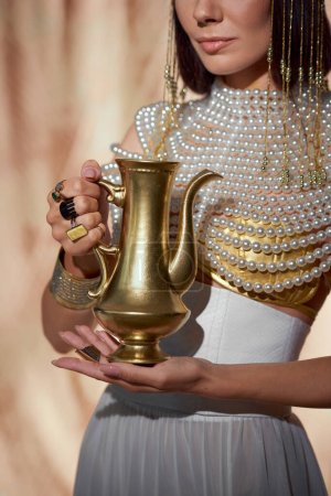 Ausgeschnittene Ansicht einer stilvollen Frau im ägyptischen Look mit goldenem Krug auf abstraktem Hintergrund