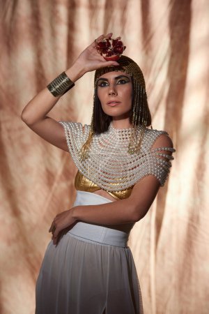 Stilvolle Frau im ägyptischen Look und kühnem Make-up mit geschnittenem Granatapfel auf abstraktem Hintergrund