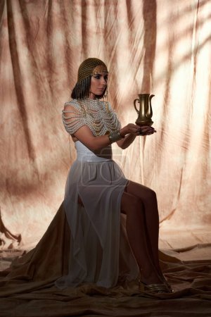 Frau in traditionellem ägyptischen Outfit hält goldenen Krug während sie auf abstraktem Hintergrund sitzt