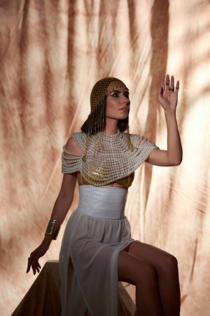 Stilvolle Frau mit ägyptischem Kopfschmuck und Perlentop posiert und sitzt auf abstraktem Hintergrund