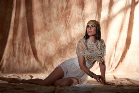 Brünette Frau in ägyptischem Look und Perlentop, die auf abstraktem Hintergrund mit Sonnenlicht wegschaut