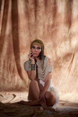 Elegante Frau mit ägyptischem Kopfschmuck und Perlenoberteil blickt in die Kamera auf abstraktem Hintergrund
